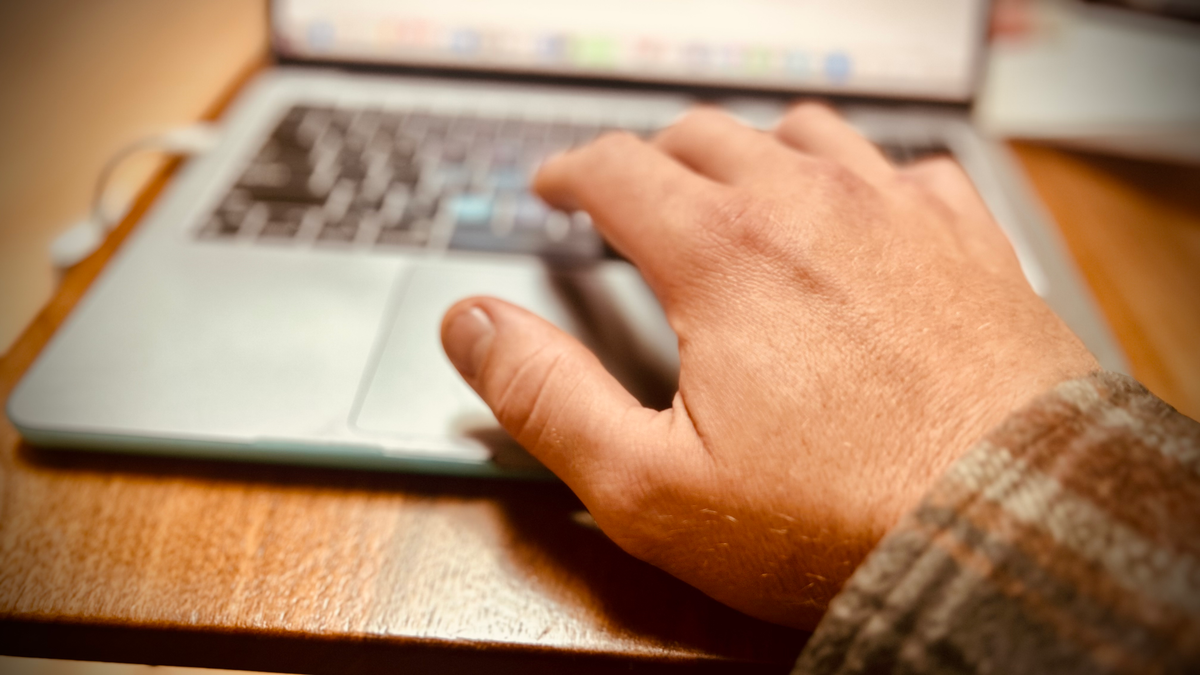 POV - my right hand reaches out to type on a macbook pro. I'm wearing brown plaid shirt. My hand is in focus. I'm working at a wood dining table. The laptop is plugged into power.